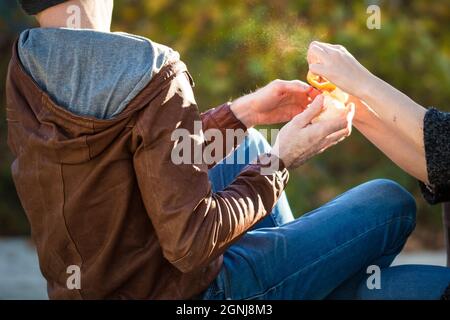 Das junge Paar spielt im Freien und schält eine Orangenfrucht. Männliche Hände in brauner Lederjacke halten Zitrus und Frau bereit, es zu fangen. Junge Familie im Freien Stockfoto