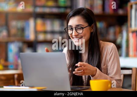 Positive arabische Frau, die eine Videokonferenz auf einem Laptop veranstalte, im Stadtcafe saß, sprach und gestikulte vor der Computer-Webcam Stockfoto