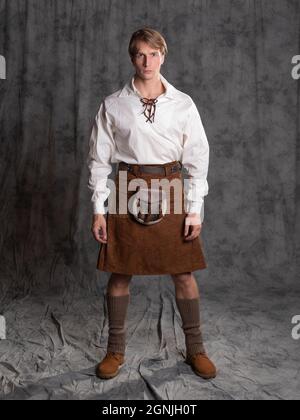 Ein junger Mann in einem Lederkilt und einer weißen Schnürbluse. Ein schottisches Foto in voller Länge im Studio auf grauem Hintergrund Stockfoto