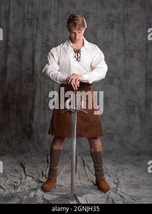 Ein junger Mann in einem Lederkilt und einer weißen Schnürbluse. Ein schottischer Ritter mit einem zweihändigen Schwert. Foto in voller Länge im Studio auf einem grauen Hintergro Stockfoto