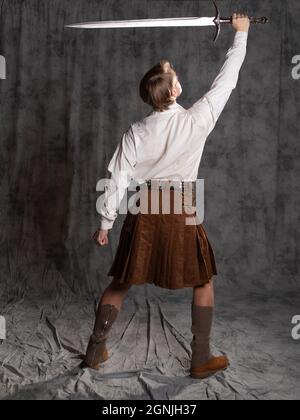 Ein junger Mann in einem Lederkilt und einer weißen Schnürbluse. Er hob die Waffe über seinen Kopf. Ein schottischer Ritter mit einem zweihändigen Schwert. Durchgehende P Stockfoto