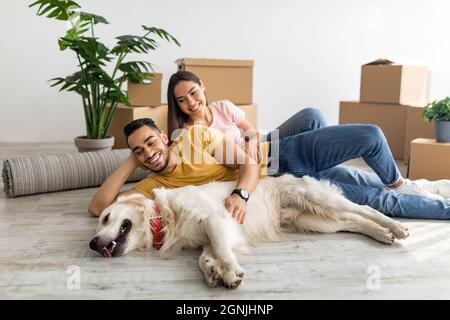 Lächelndes multirassisches Paar mit ihrem Hund, der auf dem Boden zwischen Kartons liegt, drinnen Stockfoto