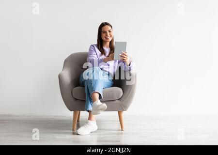 Die ganze Länge der fröhlichen jungen Frau mit Tablet-pc sitzt im Sessel, arbeitet oder studiert online gegen weiße Wand Stockfoto