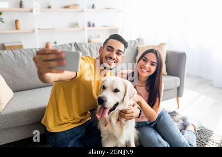 Junges liebevolles multirassisches Paar, das zu Hause Selfie mit Hund nimmt Stockfoto