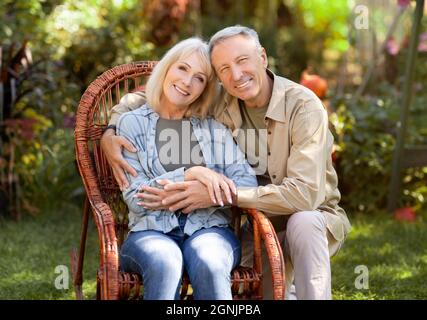 Lächelnde, liebevolle ältere Frau, die auf einem Korbstuhl sitzt, Mann umarmt Frau und hält sich die Hände Stockfoto