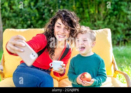 Glückliche Mutter und Sohn chillen draußen auf einer Couch in einem öffentlichen Park Garten sitzen und machen ein Selfie-Portrait auf dem Smartphone. Lächelnde Frau und Kind Stockfoto