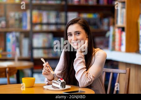 Hübsche arabische Frau, die Dessert und Kaffee genießt, während sie im Café sitzt und die Kamera anlächelt, den Kopierraum Stockfoto