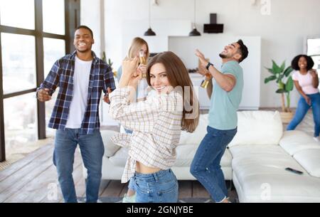 Glückliche junge Dame mit ihren vielfältigen Freunden, die zu Hause Party, Tanz und Bier trinken Stockfoto