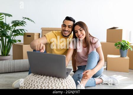 Fröhliches, vielfältiges Paar mit einem Laptop, das online nach Haushaltswaren für sein neues Zuhause sucht und am Tag des Umzugsverkehrs einen pc verwendet Stockfoto