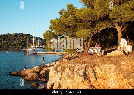 Podvrske, Murter, Kroatien - 26. August 2021: Pinien am felsigen Strand, Menschen, die Strandzubehör tragen und Boot im Sommer Sonnenuntergang festmachen Stockfoto