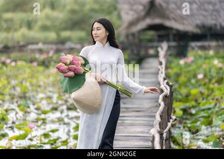 Porträt einer schönen vietnamesischen Frau mit traditionellem vietnam-Hut, der den rosa Lotus hält, der auf der Holzbrücke im großen Lotussee, vietnam, ai, läuft Stockfoto