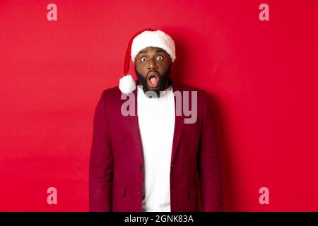 Weihnachts-, Party- und Feiertagskonzept. Schockiert Black man in santa Hut fallen Kiefer und starren auf die Kamera, stehen vor rotem Hintergrund Stockfoto