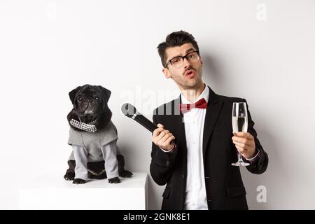 Schöner ausgefallener Mann in Gläsern, das Anheben eines Glases Champagner und das Geben des Mikrofons zu süßem Mops im Partyanzug, feiern und Spaß haben, weiß Stockfoto