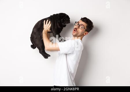 Bild eines hübschen jungen Mannes, der seinen Mops liebt. Hundebesitzer hält Welpen und lächelt glücklich an der Kamera, stehen auf weißem Hintergrund Stockfoto