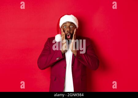 Weihnachts-, Party- und Feiertagskonzept. Schockiert Black man in santa hat Reaktion auf Neujahr Angebot, stehen vor rotem Hintergrund Stockfoto