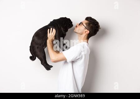 Profil von schönen jungen Mann küssen kleinen niedlichen Hund Gesicht. Hipster Kerl liebt seinen Mops, stehen über weißem Hintergrund Stockfoto