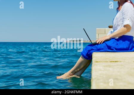 Arbeiten Sie von überall aus. Seitenansicht einer jungen Frau, Freiberuflerin im Strohhut, die am Laptop arbeitet, während sie am Strand sitzt Stockfoto