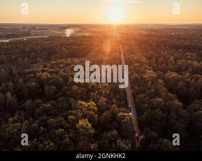 Blick von einer Höhe auf den Boden auf den Wald und die Straße im Wald. Der Sonnenuntergang und die Stadt sind im Hintergrund zu sehen. Stockfoto