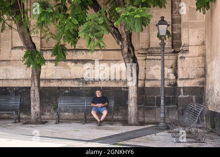 Ein Mann, der an einem heißen Sommernachmittag vor der barocken Kathedrale von Acireale aus dem 17. Jahrhundert auf der Piazza Duomo in Acireale im Schatten schläft. Catania, Sizilien, Italien Stockfoto