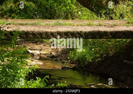 Ländliche kleine Betonbrücke über kleinen flachen Bach und felsige Landschaft in den Bergen. Verlassene Betonplattenbrücke über einen Bach. Stockfoto