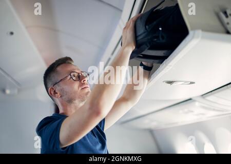 Junger Mann reist mit dem Flugzeug. Passagier, der Handgepäck in Schließfächern über den Sitzen des Flugzeugs verstaut. Stockfoto