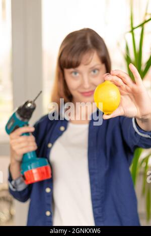 Eine junge hübsche Frau in einem blauen Hemd hält einen Schraubendreher mit einem Bohrer und einer hellen Zitrone vor dem Hintergrund eines hellen Fensters und grünen Blättern Stockfoto