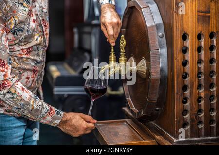 Person, die Wein aus einem Weinfass ausgießt. Reifer Sommelier im Keller des Weinguts gießt Rotwein in langstielige Weingläser. Stockfoto