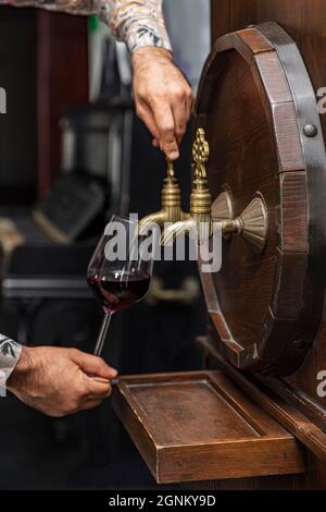 Person, die Wein aus einem Weinfass ausgießt. Reifer Sommelier im Keller des Weinguts gießt Rotwein in langstielige Weingläser. Stockfoto
