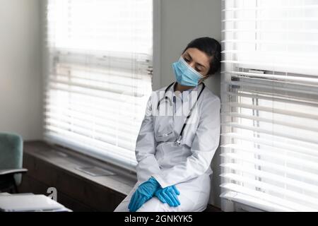 Müde indische Arzt in Maske und Handschuhe an der Wand gelehnt Stockfoto