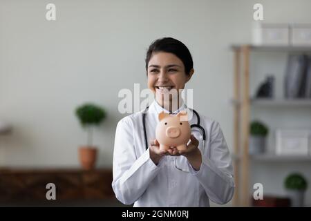 Glückliche indische junge Ärztin, die Sparschwein vor der Kamera zeigt Stockfoto