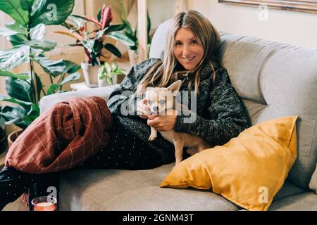 Junge Frau mit Karo und Kissen sitzt zu Hause auf der Couch, trägt bequeme Kleidung und spielt mit ihrem chihuahua Hund. Herbst Herbst und Winter Stockfoto
