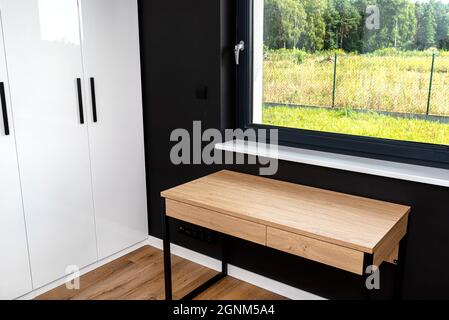 Ein Holztisch unter dem Fenster in einem modernen Jungen Zimmer in schwarz-weißen Farben, mit Vinyl-Platten auf dem Boden, großen Kleiderschrank sichtbar. Stockfoto