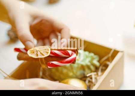 Die Hand der Frau hält Karamellrohr, getrocknete Orangenscheibe und Zimtstange. Handgefertigtes Weihnachtsdekor. Überraschung im Geschenkkarton auf dem Hintergrund. Neujahrsstimmung. Festliche Atmosphäre. Stockfoto