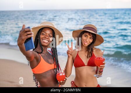 Fröhliche junge, multirassische Frauen in Strandkleidung genießen Drinks und machen Selfie auf dem Smartphone, während sie im Sommer am Sandstrand gegen das Meer stehen Stockfoto