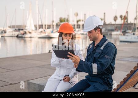 Junge männliche und weibliche Hafenmitarbeiter in Schutzhelmen und Uniform analysieren Daten auf Tablets, während sie während eines Treffens am Ufer über Arbeitsfragen diskutieren Stockfoto