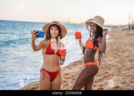 Fröhliche, unbeschwerte junge, multirassische Freundinnen in Bikinis und Sonnenhüten, die Selfie auf dem Smartphone machen und erfrischende Getränke genießen, während sie sich gemeinsam am Sandstrand ausruhen Stockfoto