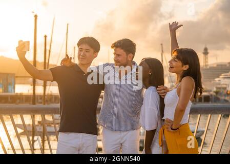 Positive junge, vielfältige Freunde umarmen und lachen beim Selfie auf dem Handy am Kai während der Sommerferien zusammen am Meer Stockfoto