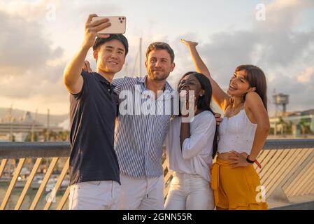 Eine Gruppe von begeisterten, tausendjährigen multirassischen Freunden in legerer Kleidung, die sich am Ufer treffen und Selfie auf dem Smartphone machen, während sie das Sommerwochenende gemeinsam in der Stadt genießen Stockfoto