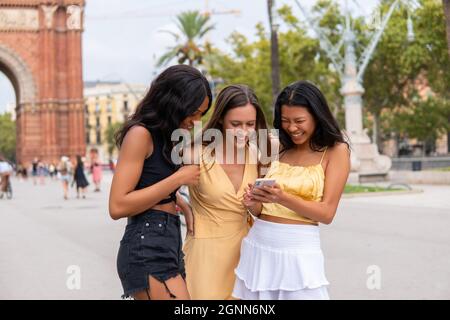 Junge asiatische Frau, die Fotos auf dem Smartphone zeigt, um verschiedene Freundinnen in stilvoller Sommerkleidung zu lachen, während sie während der Sommerferien den historischen Ort in Barcelona besucht Stockfoto