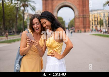 Junge multirassische Freundinnen in trendigen gelben Outfits, die auf den Handybildschirm schauen und lachen, während sie Fotos schauen, nachdem sie auf der Sommerreise auf der Straße von Barcelona City zusammen waren Stockfoto