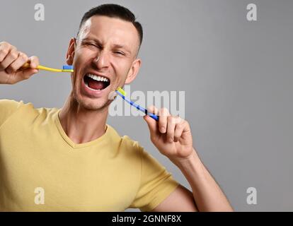 Laut schreiender Erwachsener in gelbem T-Shirt hält zwei Zahnbürsten an seinem Mund. Gesunder Lebensstil Stockfoto
