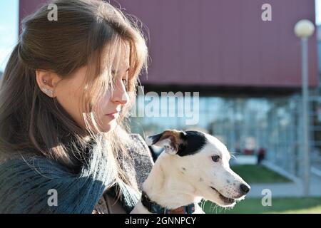 Junge Frau in einem Mantel umarmt ihren Hund, während sie auf einer Bank in einer Stadt sitzt. jack russel Terrier auf den Knien der Frau schaut zur Seite. Tierpflege, zusammen gehen und sich im Freien ausruhen Stockfoto