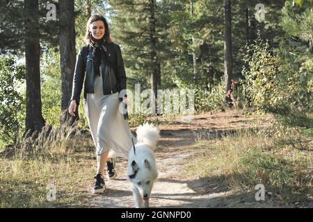Junge Frau, die mit einem Hund im Wald spazieren geht. Sonniger Herbsttag. Lächelnde Frau, die Kleid und Jacke trägt und die Kamera anschaut und Spaß mit dem weißen jakutian laika hat, während sie den Pass entlang geht. Stockfoto