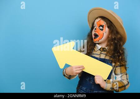 Portrait von kleinen Mädchen Kind mit Halloween Make-up-Maske, trägt Hut und Hemd, hält gelben Pfeil, zeigt weg auf leeren Raum für Promotion-Inhalte Stockfoto