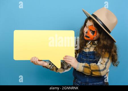 Entzückendes kleines Mädchen mit Halloween Make-up Maske, trägt braunen Hut mit großen gelben leeren Sprechblase, posiert isoliert über blauen Hintergrund Wand Stockfoto