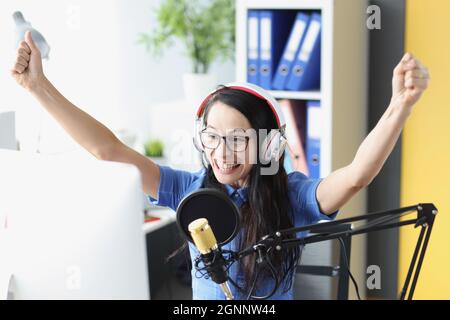 Freudig lächelnde Frau hebt begeistert die Hände in Kopfhörer und Mikrofon und schaut auf den Computermonitor Stockfoto