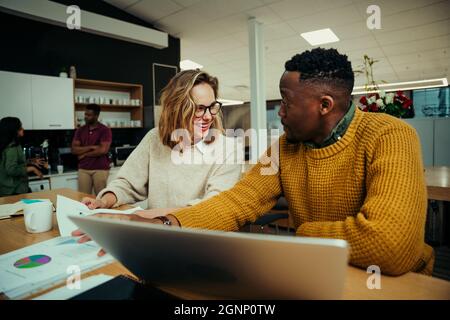 Geschäftsfrau lacht und Mann, während sie Informationen auf einem Laptop recherchiert und im Büro über Dokumente diskutiert Stockfoto