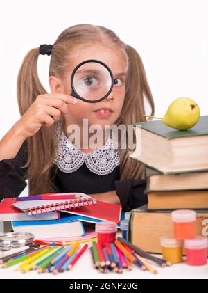 Kleines Mädchen, blond in einem Schulkleid, schaut durch eine Lupe auf weißem Hintergrund Stockfoto