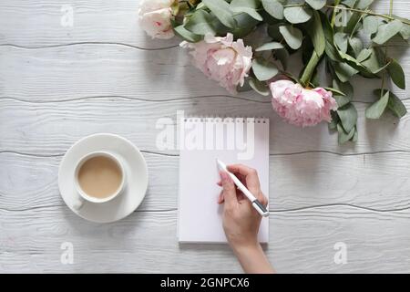 Die Hand einer Frau schreibt in ein Notizbuch. Tisch mit Pfingstrosen und einer Tasse Kaffee von oben. Stockfoto