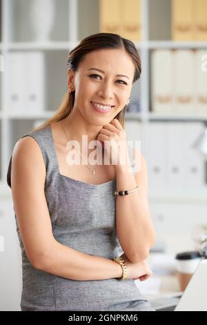 Porträt einer lächelnden jungen vietnamesischen Geschäftsfrau in Juwelen, die den Hals mit Ordnern im Büro gegen die Regale berührte Stockfoto